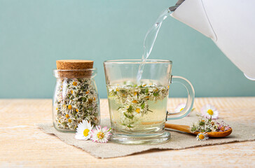 Pouring boiling water from electric kettle to a herbal tea cup. Dried herbal medicinal plant Common Daisy, also known as Bellis Perennis. Dry flower blossoms in glass jar and and herbal tea in glass.