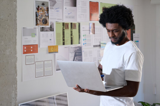 Creative Professional Researching with Laptop. An absorbed creative professional conducts research on his laptop, standing against a backdrop of project plans in a dynamic office environment.