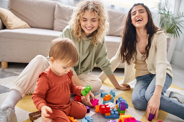 Naklejka premium cheerful beautiful lesbian couple playing with their toddler daughter at home, modern parenting