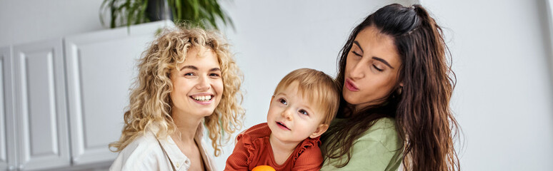 merry pretty lgbt couple having fun together with their cute baby girl holding tangerines, banner