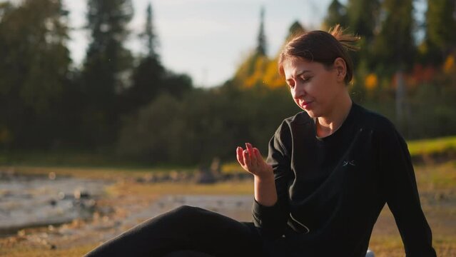 Woman resting on riverbank near forest. Depressed lady touches stones in hands alone at sunset. Devastated state and female apathy towards life