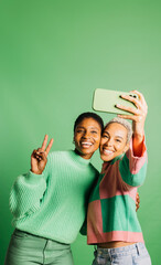 Two young happy women taking a selfie in front of a green background in a studio. Women taking a selfie in a studio.
