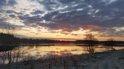 Obraz premium The sun rises from behind the forest on the far side of the lake and brightly colors the cloudy sky. Clouds and trees are reflected in the water. The grass on the shore is covered with frost