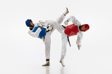 Dynamic image of young men, taekwondo athletes in kimono and helmets training isolated over white background. Concept of martial arts, combat sport, competition, action, strength, education - obrazy, fototapety, plakaty