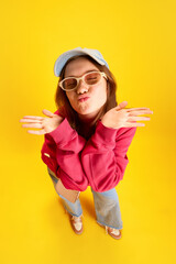 Top view. Sweet and cute funny young girl, student grimacing, stretched out her lips and sends kiss to camera against yellow background. Concept of fashion and style, traveling, culture, fun and joy.