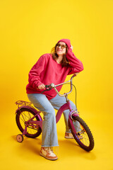 Obraz na płótnie Canvas Happy cheerful girl sitting on pink bicycle dressed trendy oversized clothes against sunny yellow background. Concept of fashion and style, hobby, traveling, culture, fun and joy.