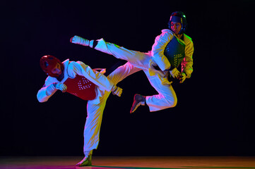 Dynamic image of young men, taekwondo athletes in kimono and helmets training against black background in neon light. Concept of martial arts, combat sport, competition, action, strength