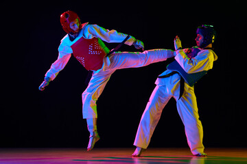 Dynamic image of young men, taekwondo athletes in kimono and helmets training against black background in neon light. Concept of martial arts, combat sport, competition, action, strength