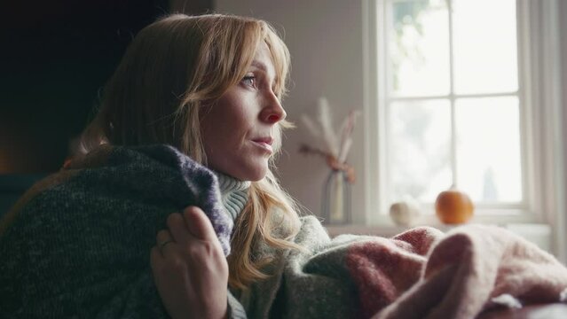 Head and shoulders shot of unhappy woman wearing cosy jumper and blanket sitting on sofa at home looking thoughtfully out of window - shot in slow motion