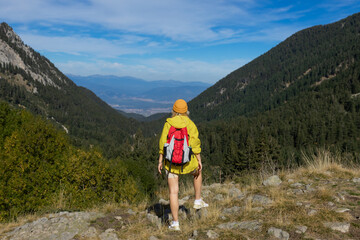 Fototapeta premium A girl on top of a mountain with green grass looks at a beautiful mountain valley. Landscape with a sporty young woman in a yellow raincoat. Travel and tourism. Hiking