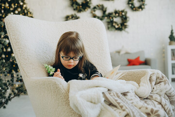 A little girl with Down syndrome reads a book while sitting in a white chair on Christmas Eve....