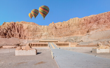 Hot air balloon flying over Hatshepsut Temple at sunrise in Valley of the Kings and red cliffs...