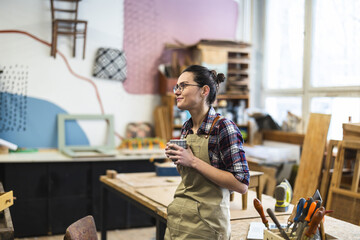 Portrait of a young female carpenter holding a cup of coffee in her workshop