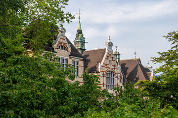 Zurich, Switzerland. View of the decorated facade of an ancient building with a clock tower through the green foliage of trees.