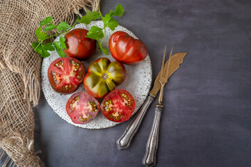 Fresh red ripe tomatoes on wooden cutting board with parsley, dark rustic background. Cut tomatoes with fork and knife. Top view, flat plan, copy space. - obrazy, fototapety, plakaty