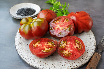 Fresh red ripe tomatoes on a wooden cutting board with black Thursday salt, dark rustic background. Tomatoes being cut with fork and knife