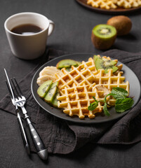 A freshly prepared waffles with kiwi and banana slices on a gray plate with a cup of coffee and a napkin on a dark background.