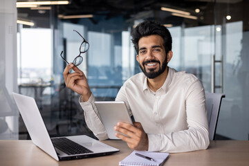 Portrait of a smiling young Indian man working in the office, sitting at a desk. holds a tablet and...