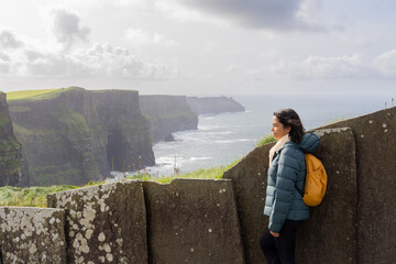 Latin woman with backpack enjoying a beautiful seascape from the top of the cliffs in Ireland.