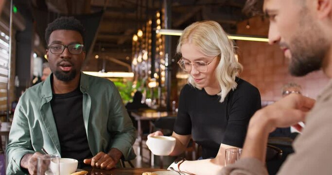 Camera view of mixed-raced friends sitting together at table in cafe. Actively speaking with each other and happily smiling. Having positive conversation. People resting in bar at weekend.