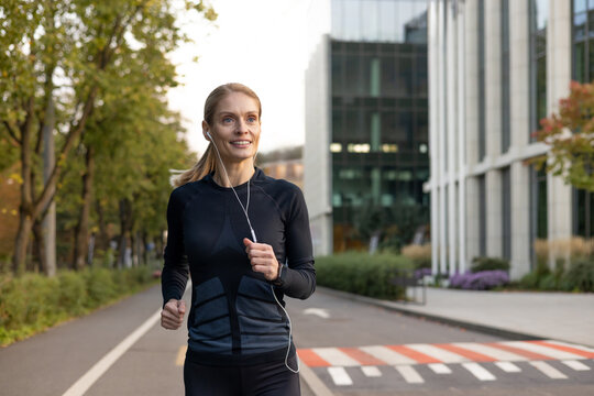 Mature experienced adult woman running in headphones in tracksuit outside modern building near trees, happy with workout sportswoman smiling active lifestyle.
