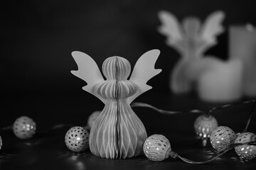 DIY paper angel on a dark background. Religious symbols, festive Christmas and New Year decor, garland lights. Black and white artistic low key photo.
