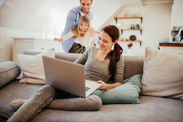 Mother working on a laptop on the couch while father and daughter play in the background at home