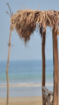 Thatched wooden huts sway in the wind by the beach with sand and choppy sea in the background during the day.