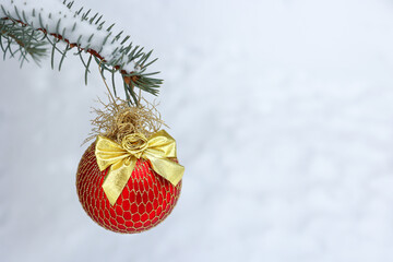 A red decoration with a golden bow of the Christmas ball hangs on a branch of a fir tree on the street, in a park covered with white snow.