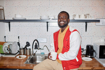 Waist up portrait of smiling Black man wearing varsity jacket smiling at camera in kitchen, with...