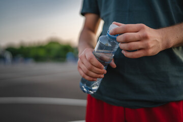 caucasian man teenager open plastic bottle of water outdoor in sunny day drink while hold basketball
