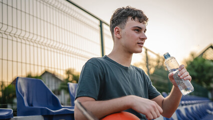 caucasian man teenager open plastic bottle of water outdoor in sunny day drink while hold basketball - obrazy, fototapety, plakaty