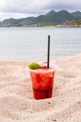 A Cup of Strawberry Daiquiri Served with Maraschino Cherry and Wedge of Lime Sitting on a Sandy Beach