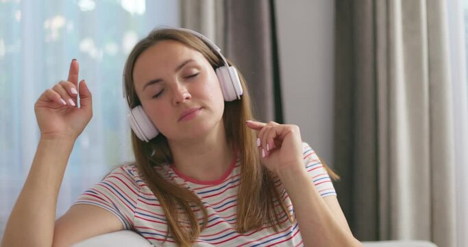 A young Caucasian woman wearing wireless headphones sits on a couch at home