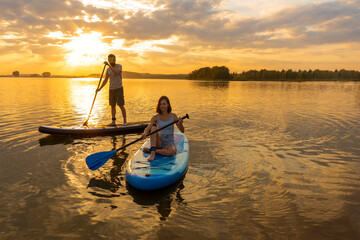 Happy couple paddle boarding at lake during sunset together with pug dog. Concept of active family...