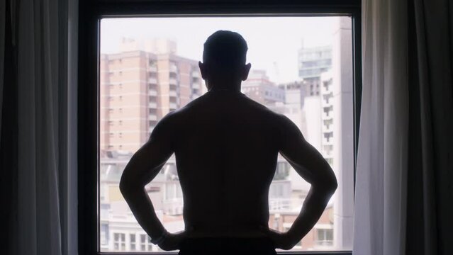 Shirtless man standing in front of window hands on hips in hotel room medium shot 24mm Low Exposure