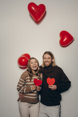 Smiling loving couple holding heart shape Valentines cards with balloons flying around