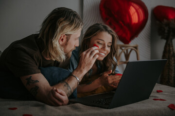 Happy loving couple shopping online while lying in bed and using laptop with heart shape balloons 