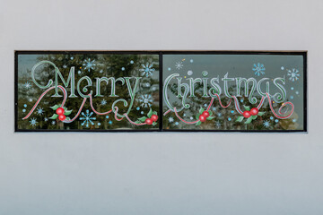 Merry Christmas is painted with colorful ink markers on glass windows with black metal trim on a white exterior wall of a building. A Christmas garland with snowflakes surrounds the grey font.