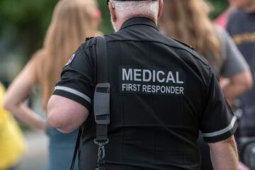 A close-up of a Caucasian male emergency health medical first responder or paramedic wearing a black uniform with grey letters. The ambulance attendant is wearing a short-sleeve shirt with lettering. - obrazy, fototapety, plakaty