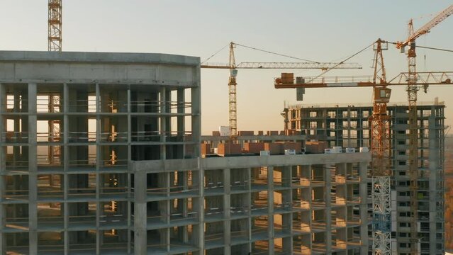 Aerial side view of concrete skyscrapers under construction with tower cranes at sunset
