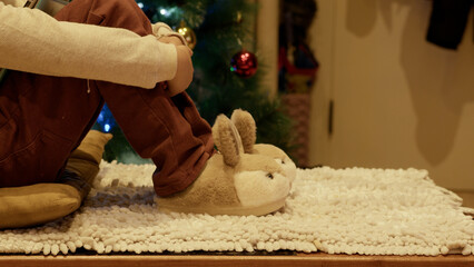 Cute boy sitting near the Christmas tree and playing with his new bunny slippers. High quality photo