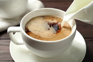 Pouring milk into cup of hot coffee on wooden table, closeup