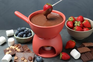 Dipping fresh strawberry in fondue pot with melted chocolate at grey table