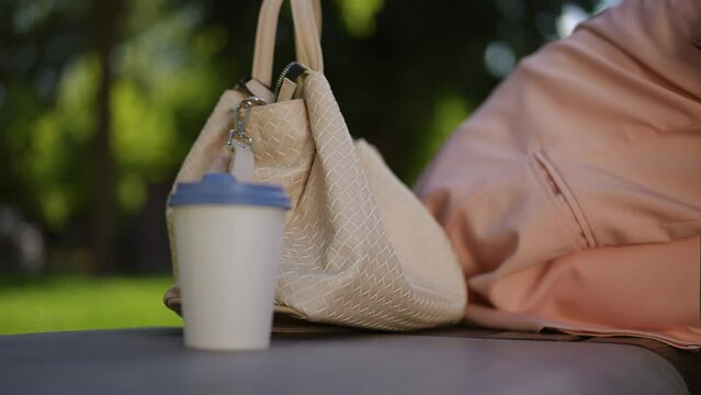 A woman sits on a park bench and places her leather bag next to her blue closed-toe sandals. The woman takes her sandals with her hand to put them on. Close-up. Body part.