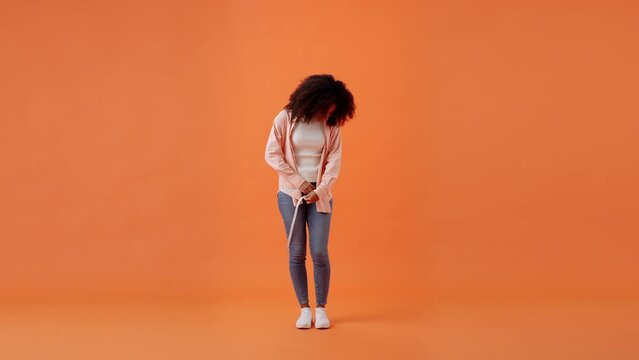 Upset African American woman measures hip girth at orange wall
