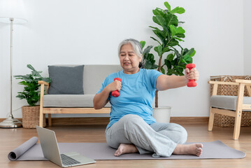 Asian elderly woman doing exercise at home by stretching the arm muscles and using a dumbbell as an...