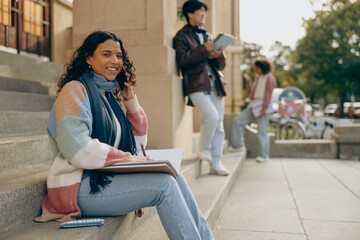 Young female student talking phone sitting near university campus on friends background