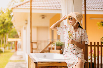 half body photo Asian female tourists come for a New Year's holiday. Looking ahead, wearing a dress, hat, holding a bouquet of flowers, glasses, sitting and relaxing in front of the villa room.