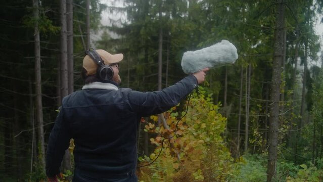 Caucasian man wearing headphones records sounds of nature for movie in coniferous wood using professional audio equipment. Sound technician works with furry windjammer microphone outdoors. Filmmaking.